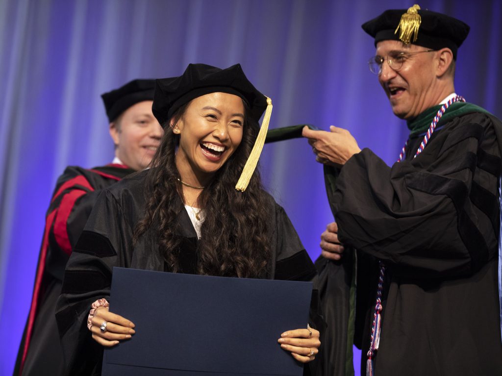MD program graduate Lisa Ho, wearing commencement robe and cap, smiles while being hooded.