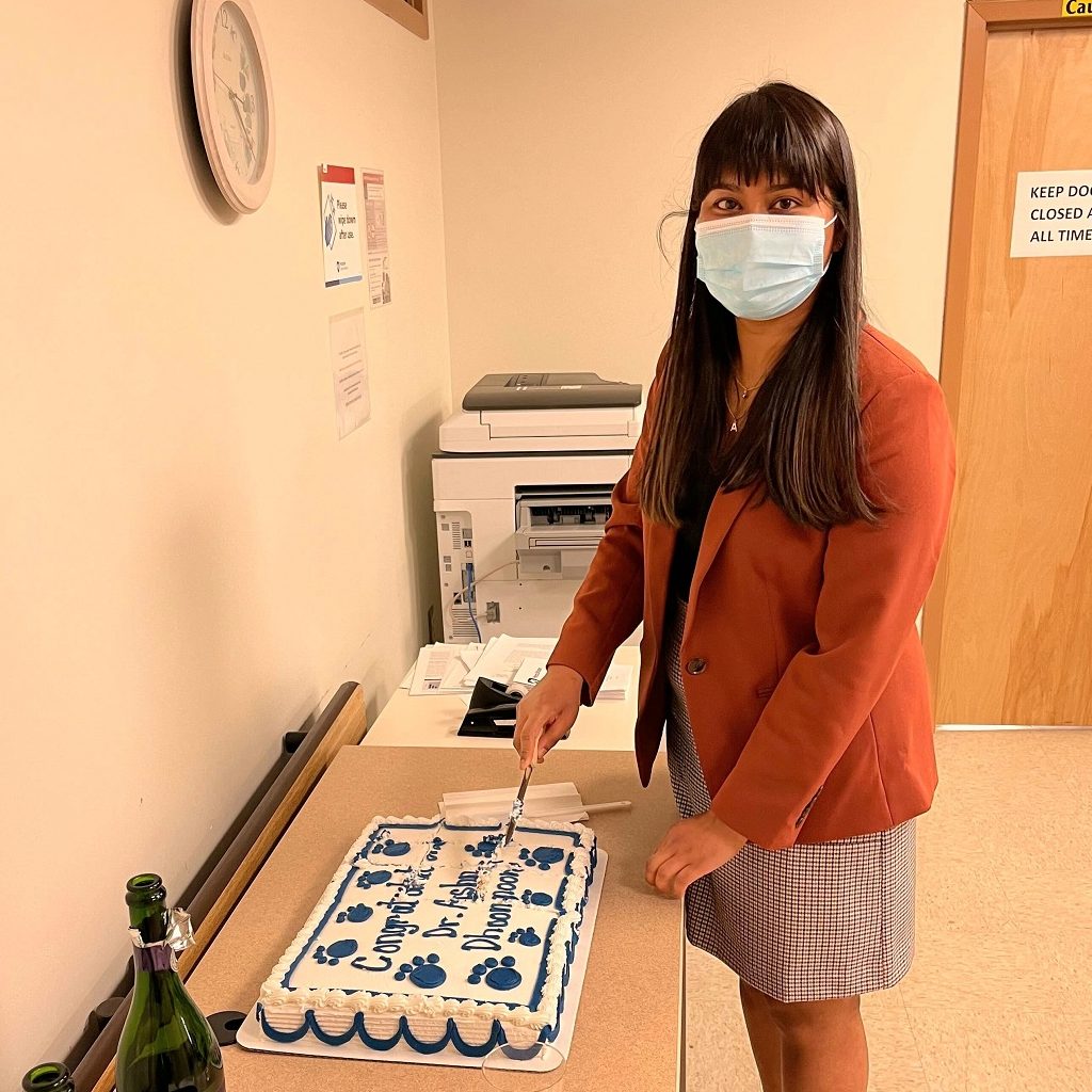 Ashna Dhoonmoon, wearing a mask, stands while cutting a cake to celebrate the defense of her dissertation.