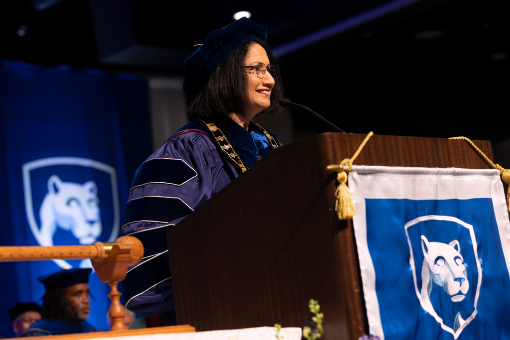 Penn State President Neeli Bendapudi looks off to the right and smiles while speaking behind a lectern with a Penn State shield banner on the front of it