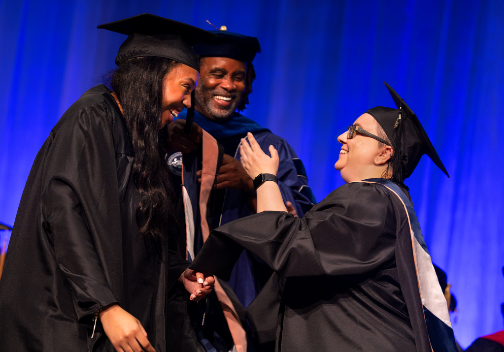 A graduate student to the left looks and smiles at a faculty member to the right as she prepares to be hooded; another faculty member smiles in the center
