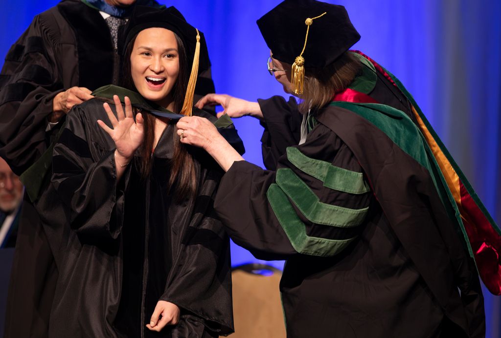 A medical student smiles a waves while being hooded by a faculty member