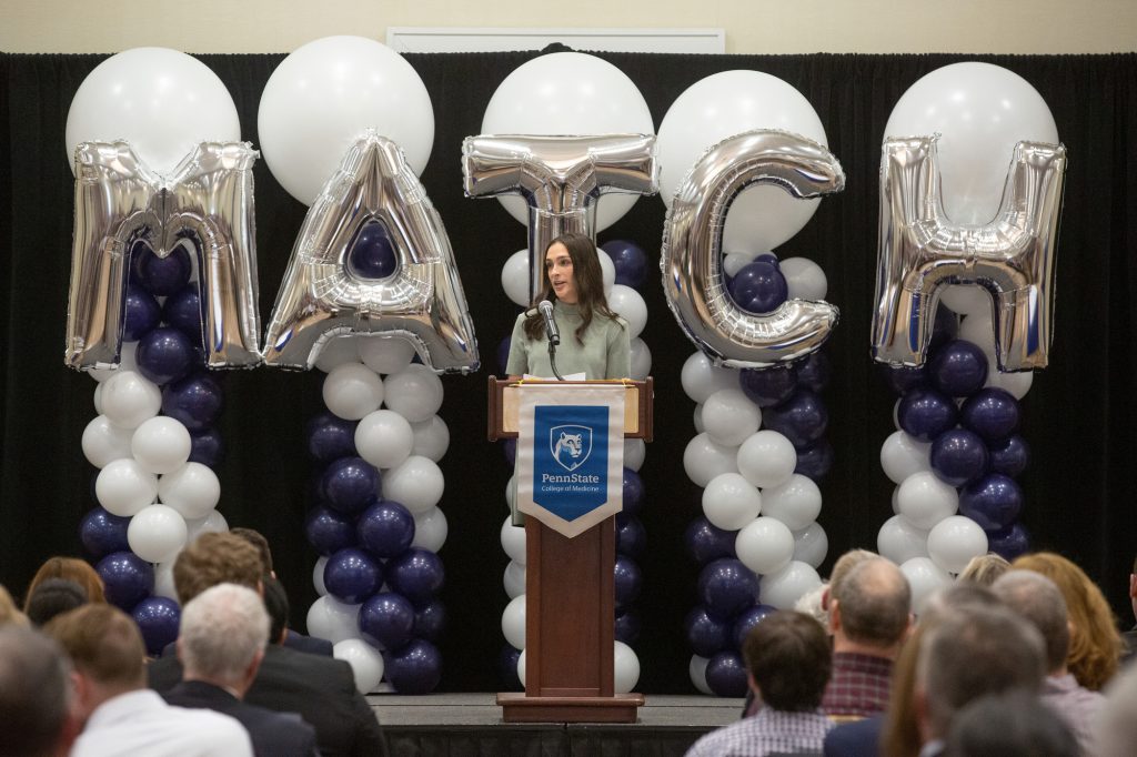 Kelly Kons speaks from a lectern with a Penn State College of Medicine banner, with balloons including balloons spelling MATCH behind her.