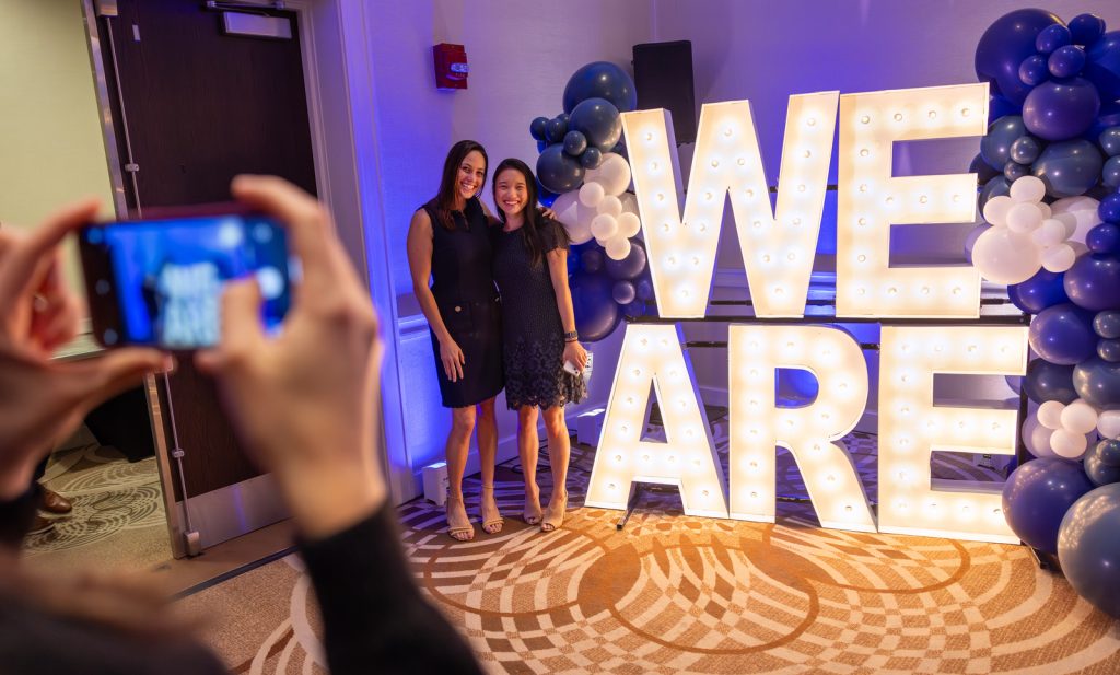 Students pose next to a large, lighted 'WE ARE' while someone takes a photo with a phone in the foreground