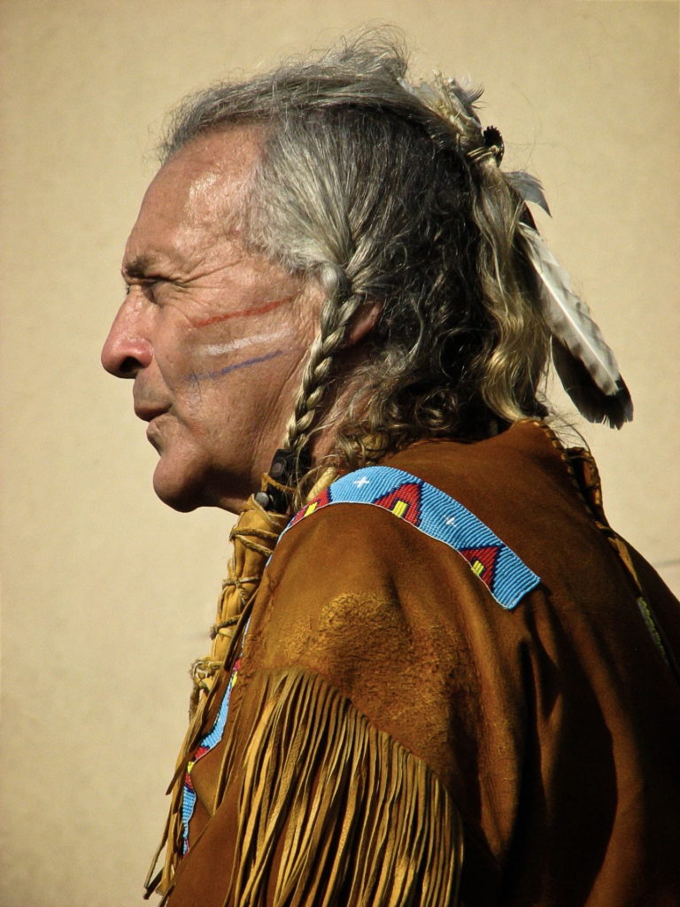 A Native American man in his 50s or 60s is seen in traditional Native dress and braided hair, in a side profile. He looks serious. The photo is by Marcia Riegle and appears in the 2020 edition of Wild Onions, Penn State College of Medicine's art and literary journal.