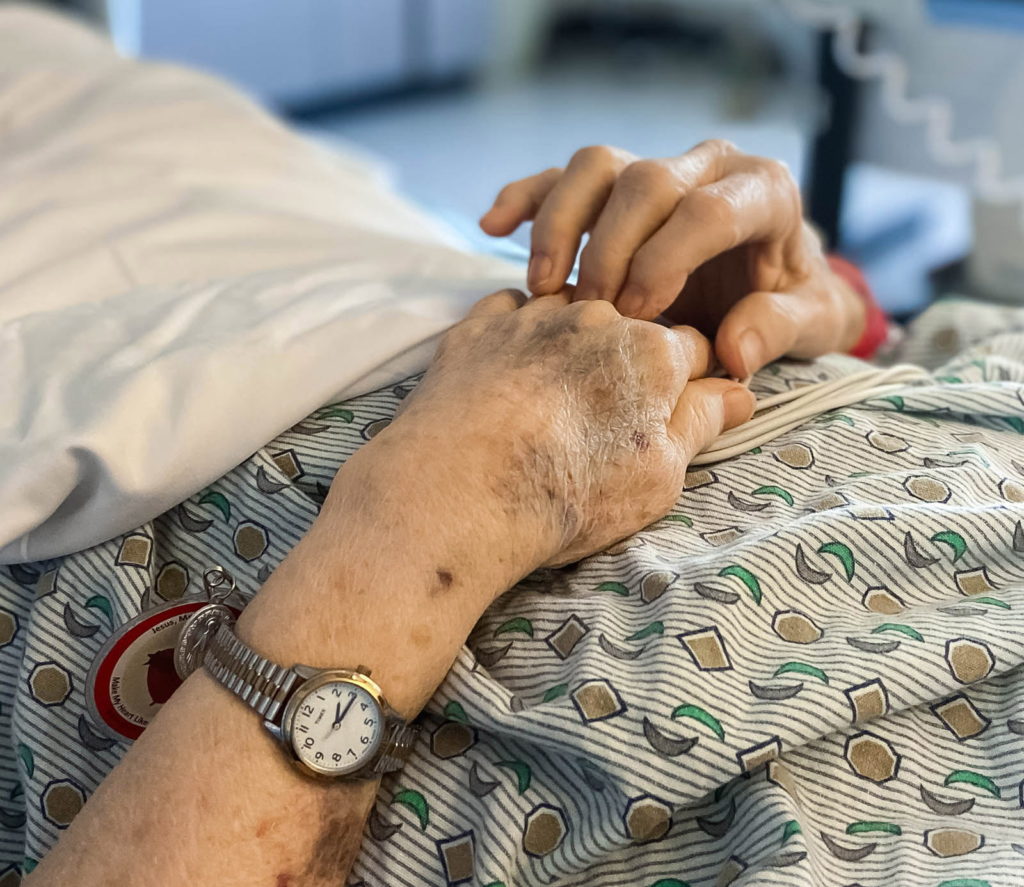 A closeup image shows the hands of a person in a hospital gown. The person's watch and a sheet are visible, but the person's face is not. The photo is by Mary Mager and appears in the 2020 edition of Wild Onions, Penn State College of Medicine's art and literary journal.