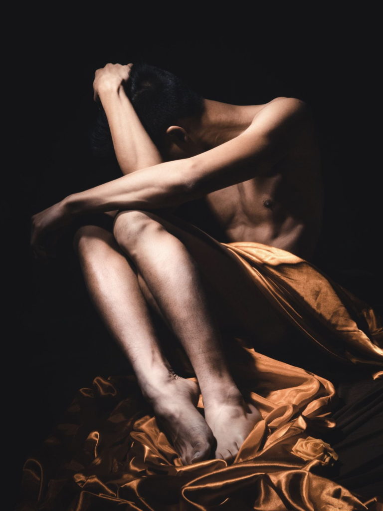 A person is seen seated, draped only in a bright cloth but otherwise naked. The background is dark and the person's arm is covering their face. The photo is by Paul Nguyen, taken in collaboration with Jacob Ritts, and appears in the 2020 edition of Wild Onions, Penn State College of Medicine's art and literary journal.